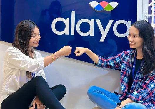 Two female colleagues, beaming with happiness, poised for a friendly fist bump while seated on the floor in front of logo of Alaya 