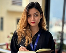 A young female in a black shirt looking straight at the camera, holding a pen and notebook in her hand. 

