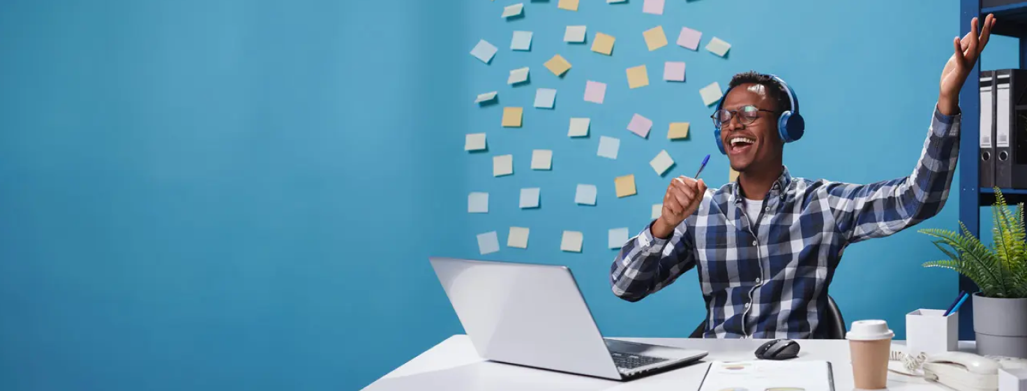 A guy sitting at a desk with a laptop and a coffee cup. He has headphones on and is holding a pen as a mic and singing happily.