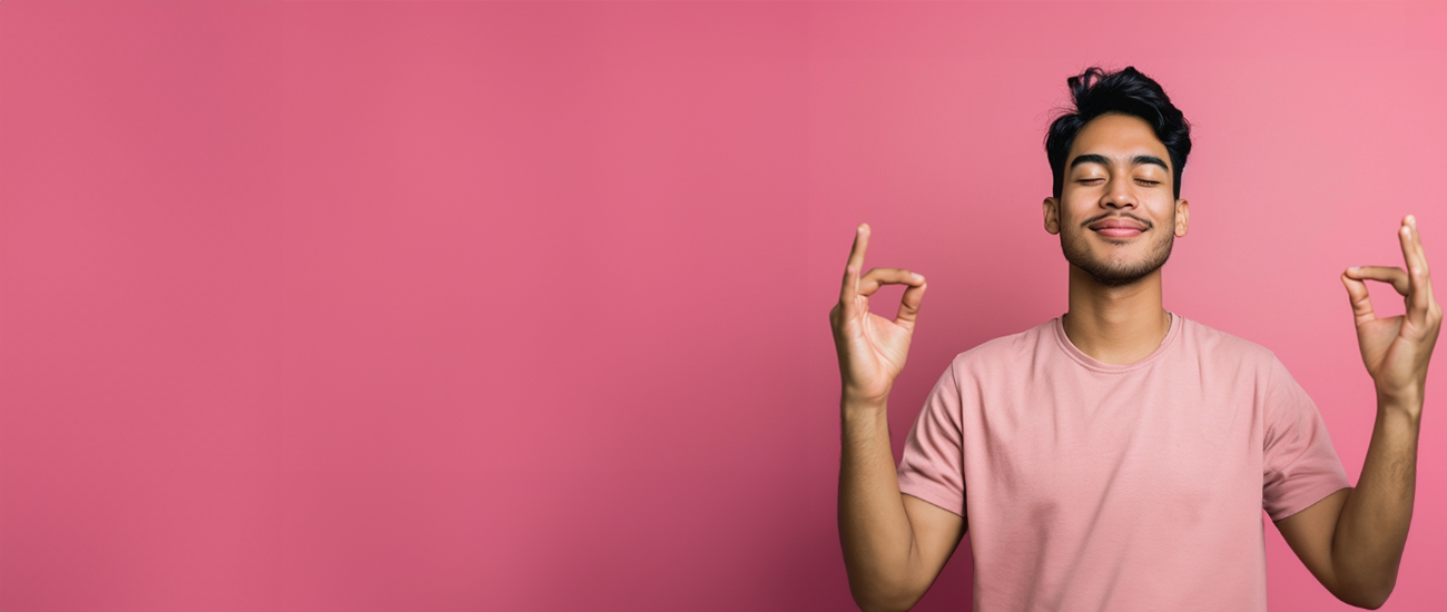 A man in a pink shirt against a pink background, holding their hands in a meditation pose, demonstrating Employee Wellness Programs.